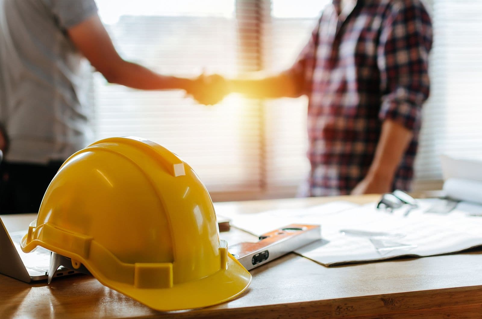 A yellow safety helmet sits on a desk with blueprints while two people shake hands in the background.