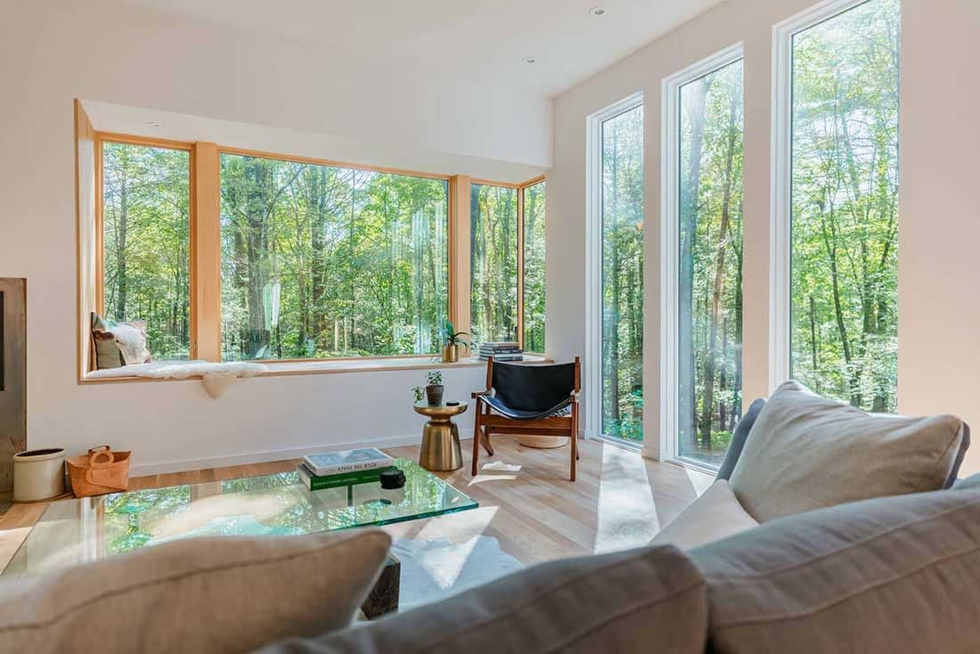 A living room with large bay and picture windows looking out over some woods.